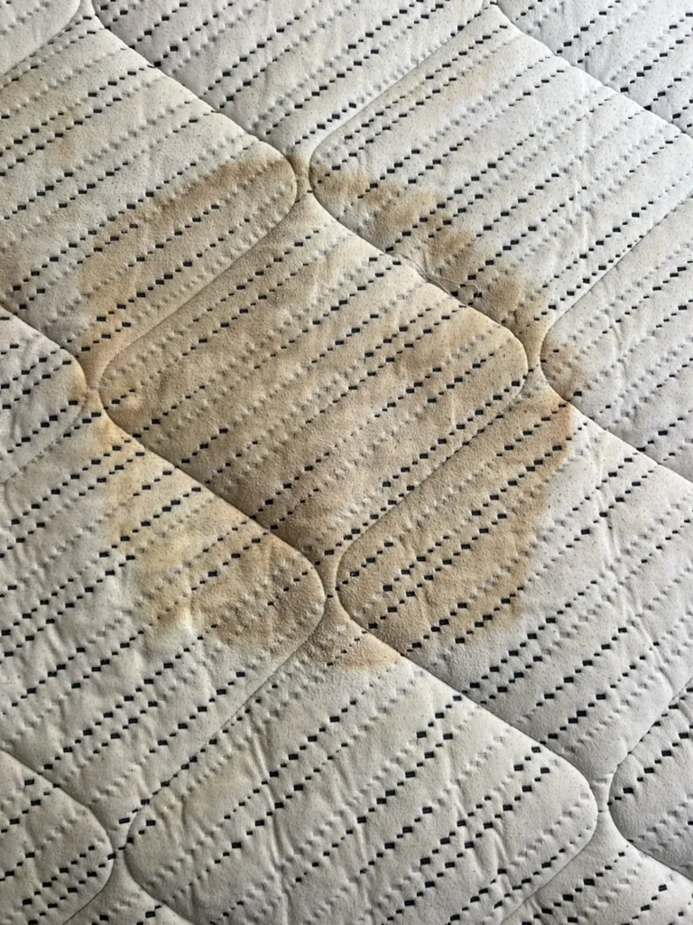 Mattress fabric showing widespread brownish staining and discolouration before professional cleaning